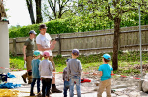 Erwachsener und Kinder tragen Kappen und arbeiten gemeinsam an der Umgestaltung des Außengeländes einer Kindertagesstätte in einen naturnahen Spielraum, daneben ein Baum und ein Zaun im Hintergrund